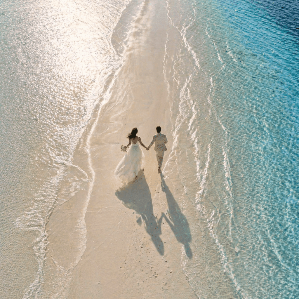 Brautpaar läuft Hand in Hand über einen Sandstrand auf den Malediven, aufgenommen aus der Vogelperspektive mit türkisblauem Meer