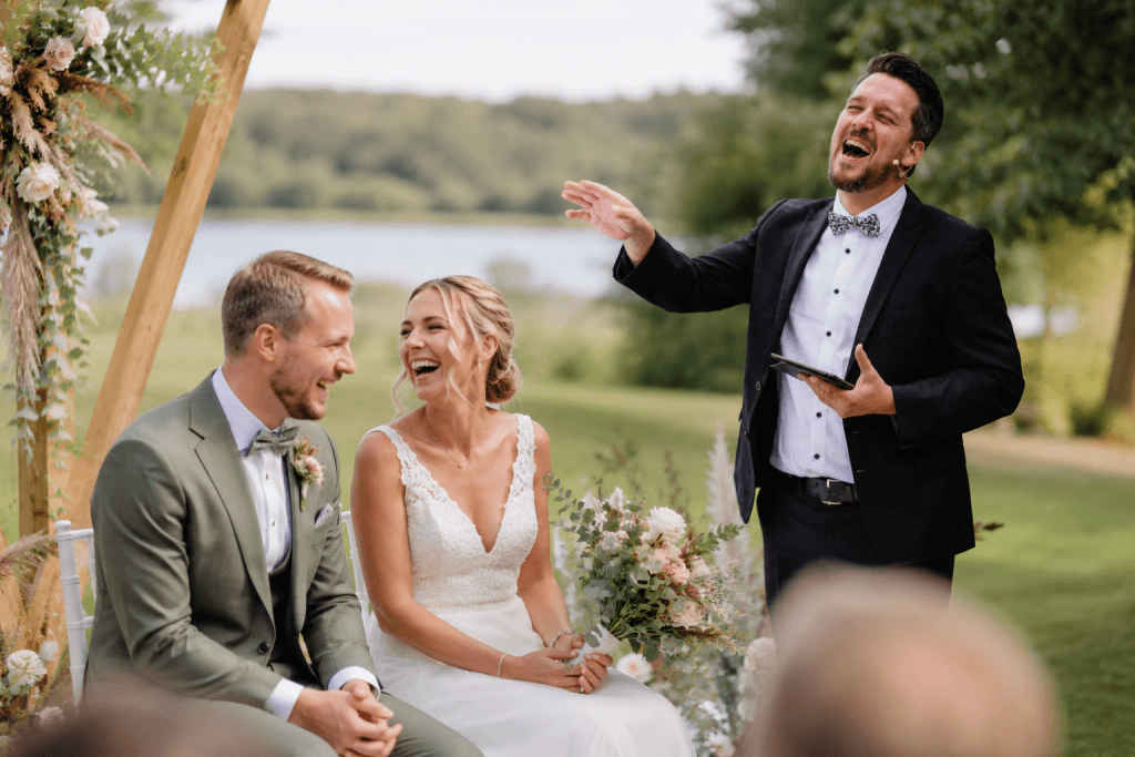 Lachendes Brautpaar bei freier Trauung im Grünen. Trauredner spricht emotional und humorvoll während der Hochzeitszeremonie am See.