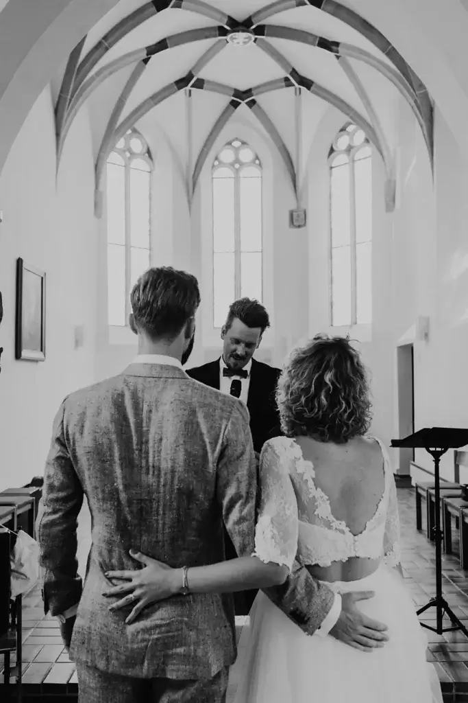 Brautpaar steht bei emotionaler kirchlicher Trauung vor einem Trauredner in einer hellen Kapelle. Moderne Hochzeit mit persönlicher Atmosphäre.
