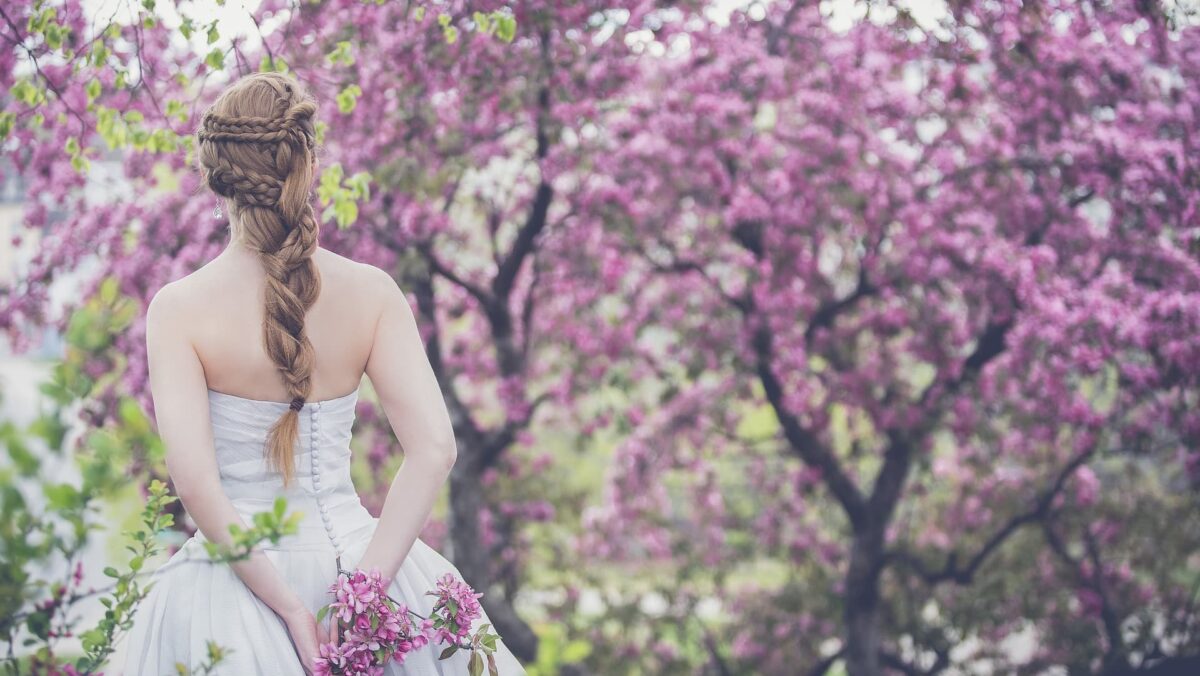 Eine Braut ist von hinten zu sehen, wie sie in ihrem weißen Brautkleid steht. Sie hält einen Blumenstrauß in der Hand und hat einen langen Schleier, der bis zum Boden reicht. Die Szene wirkt romantisch und festlich, passend zu einem Hochzeitstag.