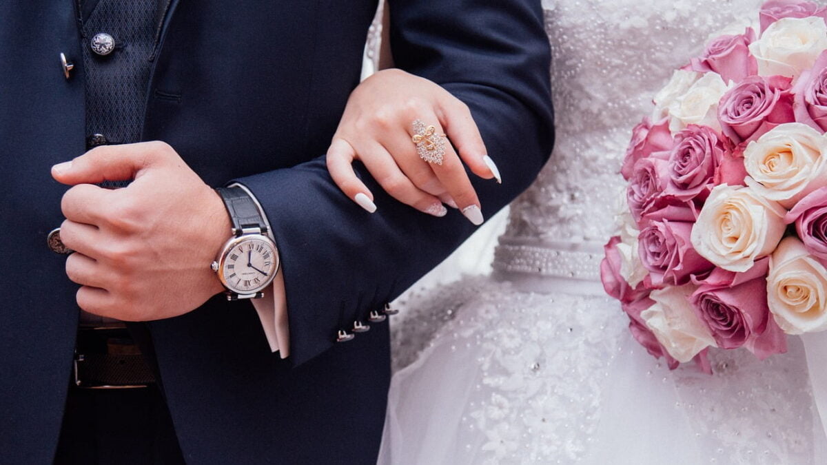 Auf dem Bild ist ein glückliches Brautpaar zu sehen, das Hand in Hand steht. Der Bräutigam trägt eine elegante Armbanduhr, die gut sichtbar ist. Beide schauen sich verliebt an und lächeln. Der Hintergrund zeigt eine idyllische Landschaft mit sanften Hügeln und einem klaren blauen Himmel, was dem Bild eine romantische und friedliche Atmosphäre verleiht.