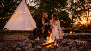 Brautpaar sitzt am Lagerfeuer im Outdoor Zentrum Lahntal und stößt mit Bier an, im Hintergrund ein beleuchtetes Zelt in der Natur